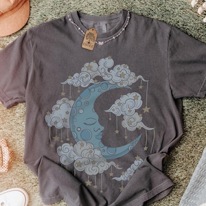 Könnte beinhalten: Ein anthrazitfarbenes Kurzarm-T-Shirt mit einem Grafikdruck eines blauen Halbmondes mit Gesicht, umgeben von Wolken und hängenden Sternen. Das T-Shirt hat einen Rundhalsausschnitt und ein braunes, rechteckiges Etikett am Kragenbereich.