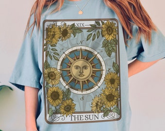 May include: A light blue t-shirt with a graphic of the The Sun tarot card. The card features a sun with a face, surrounded by sunflowers and a compass.