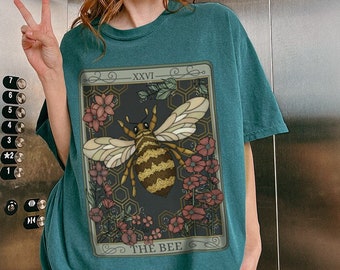 May include: A teal blue t-shirt with a graphic of a bee in a honeycomb surrounded by flowers. The graphic is in a vintage style with a black background and gold accents. The text "XXVI" and "THE BEE" are included in the graphic.