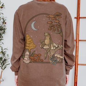 May include: A brown long-sleeved top with a fantasy illustration of a frog on a mushroom, with other mushrooms, a crescent moon, and stars. The design is framed within a dark brown arch.