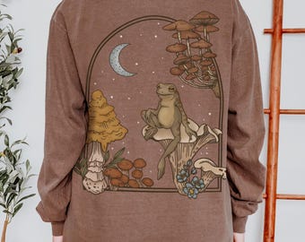 Pode incluir: Camiseta marrom de manga comprida com uma ilustração fantasiosa de um sapo sentado em um cogumelo, rodeado por outros cogumelos, uma lua crescente e estrelas. O desenho é emoldurado em um arco marrom escuro.