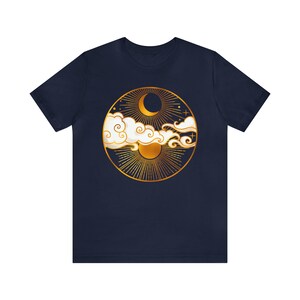 K&ouml;nnte beinhalten: Ein marineblaues T-Shirt mit einem goldenen kreisf&ouml;rmigen Design, das eine Mondsichel, die Sonne und Wolken zeigt. Das Design erinnert an eine himmlische Szene.