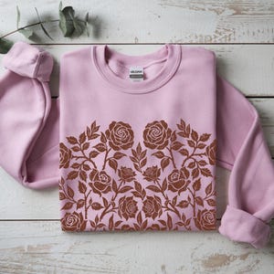 Puede incluir: Una sudadera rosa claro con un dise&ntilde;o floral marr&oacute;n en el pecho. El dise&ntilde;o presenta rosas y hojas. La sudadera est&aacute; doblada sobre una superficie de madera blanca.