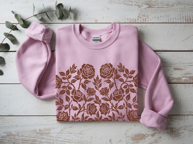 Puede incluir: Una sudadera rosa claro con un dise&ntilde;o floral marr&oacute;n en el pecho. El dise&ntilde;o presenta rosas y hojas. La sudadera est&aacute; doblada sobre una superficie de madera blanca.