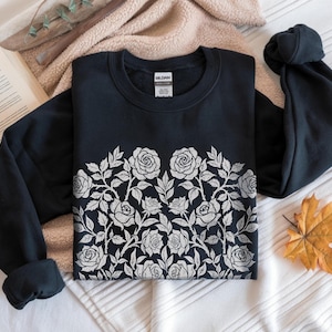 Puede incluir: Una sudadera negra con un dise&ntilde;o floral de rosas blancas en el pecho. Las rosas y las hojas est&aacute;n detalladas. La sudadera est&aacute; sobre una superficie blanca y beige con un libro y una hoja de oto&ntilde;o.