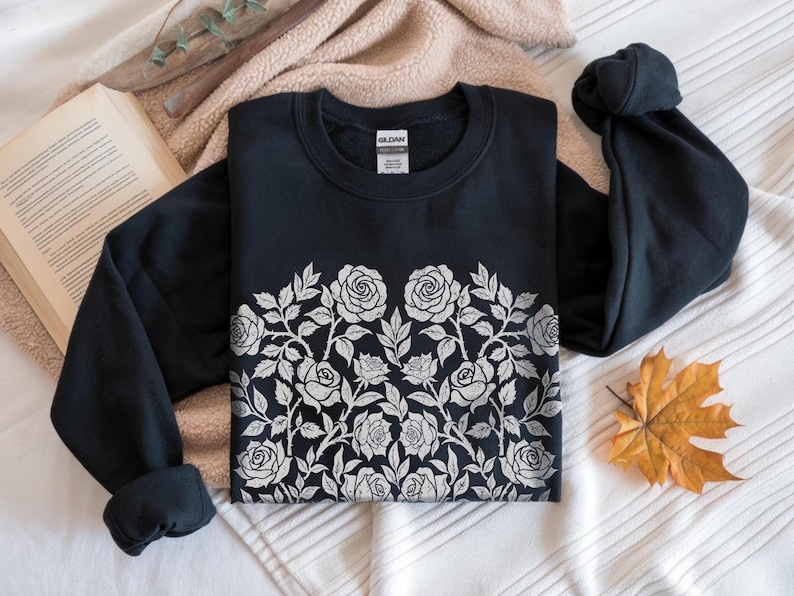 Puede incluir: Una sudadera negra con un dise&ntilde;o floral de rosas blancas en el pecho. Las rosas y las hojas est&aacute;n detalladas. La sudadera est&aacute; sobre una superficie blanca y beige con un libro y una hoja de oto&ntilde;o.