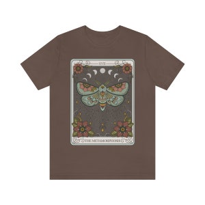 May include: A brown t-shirt with a graphic of a tarot card featuring a moth with floral accents and the text "XVIII THE METAMORPHOSIS".