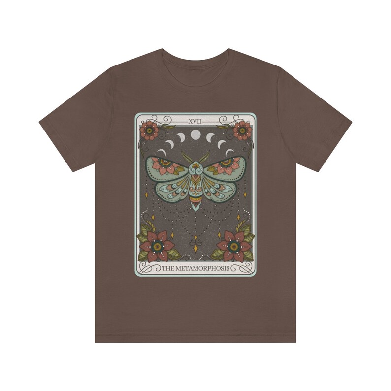 May include: A brown t-shirt with a graphic of a tarot card featuring a moth with floral accents and the text "XVIII THE METAMORPHOSIS".