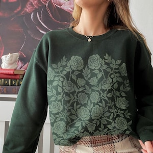 Puede incluir: Sudadera verde oscuro con un dise&ntilde;o floral de rosas en un tono verde m&aacute;s claro. La sudadera de cuello redondo tiene mangas largas y un ajuste relajado. El patr&oacute;n floral cubre la parte delantera, creando un tema bot&aacute;nico.