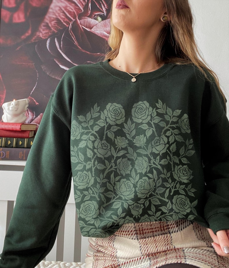 Puede incluir: Sudadera verde oscuro con un dise&ntilde;o floral de rosas en un tono verde m&aacute;s claro. La sudadera de cuello redondo tiene mangas largas y un ajuste relajado. El patr&oacute;n floral cubre la parte delantera, creando un tema bot&aacute;nico.