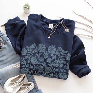 Puede incluir: Una sudadera azul marino con un dise&ntilde;o floral de rosas en tonos azules. La sudadera de cuello redondo se combina con vaqueros de lavado claro, zapatillas blancas y un collar plateado. Tambi&eacute;n se ve una peque&ntilde;a planta suculenta.