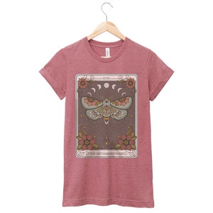 May include: A maroon t-shirt with a tarot card design featuring a moth with a starry background and the text "XVII THE METAMORPHOSIS".