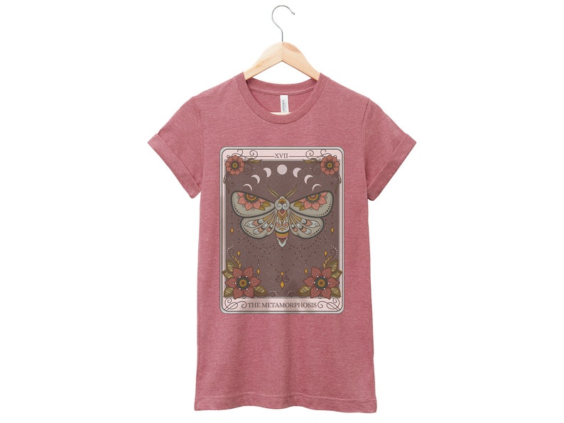 May include: A maroon t-shirt with a tarot card design featuring a moth with a starry background and the text "XVII THE METAMORPHOSIS".