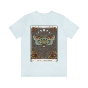 May include: A light blue t-shirt with a graphic of a tarot card featuring a moth with intricate details and the text "The Metamorphosis" on a dark brown background.