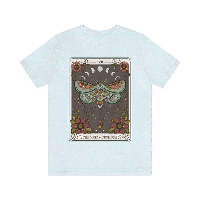 May include: A light blue t-shirt with a graphic of a tarot card featuring a moth with intricate details and the text "The Metamorphosis" on a dark brown background.