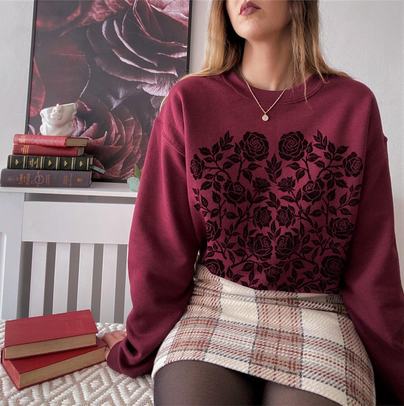 Puede incluir: Una sudadera burdeos con un dise&ntilde;o floral de rosas negras. La sudadera se combina con una falda a cuadros. La imagen tambi&eacute;n muestra una pila de libros y una obra de arte con tem&aacute;tica de rosas en el fondo.