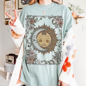 Könnte beinhalten: Ein hellblaues T-Shirt mit einem grafischen Druck einer Sonne und eines Mondes mit floralen Details. Sonne und Mond sind von einem floralen Rahmen umgeben.
