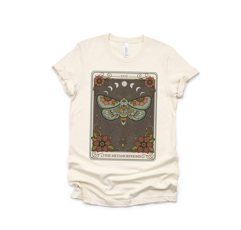 May include: A beige t-shirt with a graphic print of a tarot card featuring a moth with colorful wings and the text "The Metamorphosis" and "XVII".