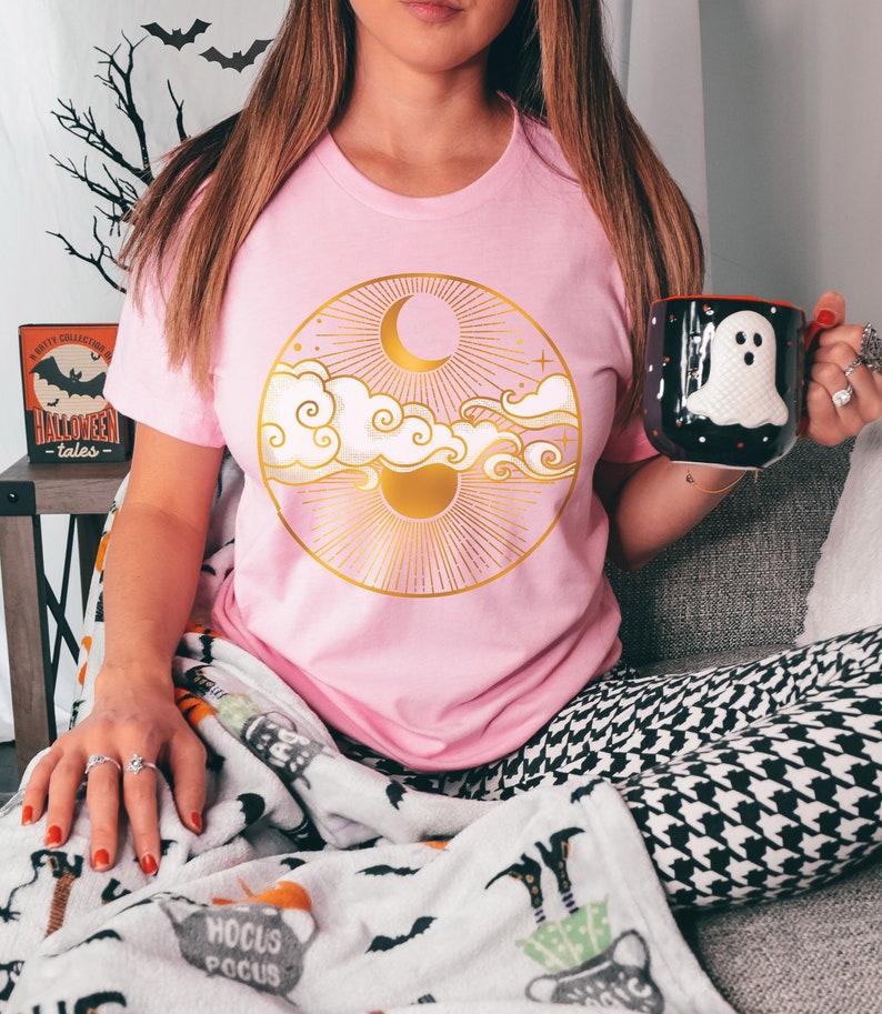 K&ouml;nnte beinhalten: Ein pinkfarbenes T-Shirt mit einem goldenen Sonnen- und Monddesign in einem Kreis. Das Design enth&auml;lt Wolken und Sterne. Das Shirt wird von einer Person getragen, die auf einem Sofa sitzt.