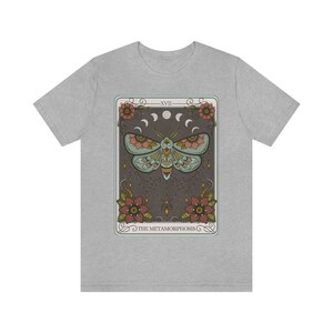 May include: A grey t-shirt with a graphic of a tarot card featuring a moth with floral accents and the text "XVIII THE METAMORPHOSIS".