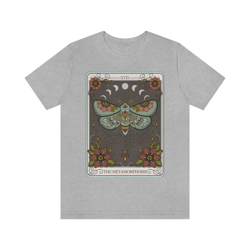 May include: A grey t-shirt with a graphic of a tarot card featuring a moth with floral accents and the text "XVIII THE METAMORPHOSIS".