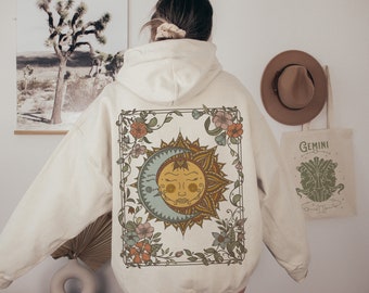May include: A cream-colored hoodie with a floral design featuring a sun and moon with a face. The design is framed with a decorative border.