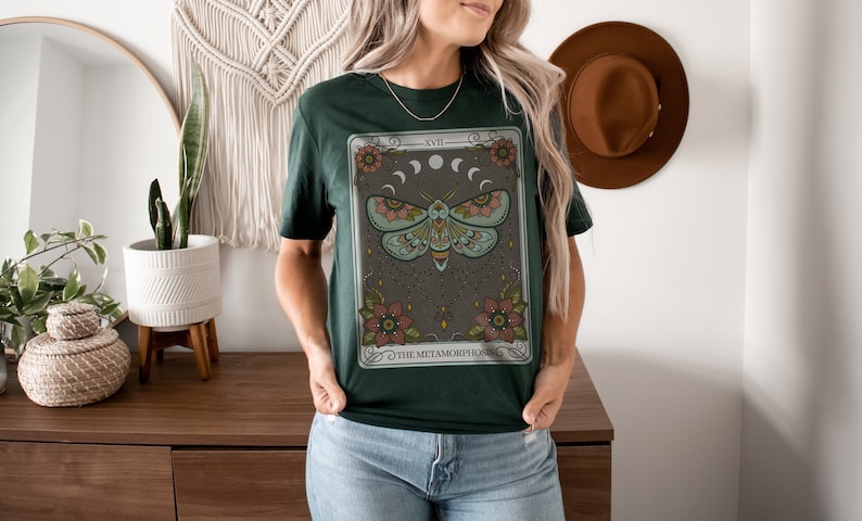 May include: A green t-shirt with a graphic of a moth and floral design. The graphic is a tarot card with the text "XVII THE METAMORPHOSIS".