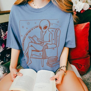 May include: A blue t-shirt featuring a graphic of an alien reading a book in a chair. The text on the shirt reads "Exploring Worlds, One Page at a Time".