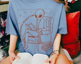 May include: A blue t-shirt with a graphic of an alien reading a book in a chair. The text on the shirt reads "Exploring Worlds, One Page at a Time".