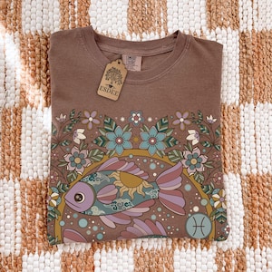 Könnte beinhalten: Braunes T-Shirt mit einem farbenfrohen Fischdesign, umgeben von Blumen und einem Sonnengrafik. Das Sternzeichen Fische ist abgebildet. Ein Etikett mit dem Text "ESDEE" ist angebracht.