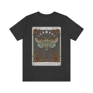 May include: A dark grey t-shirt with a graphic of a tarot card featuring a moth with floral accents and the text "XVII THE METAMORPHOSIS".