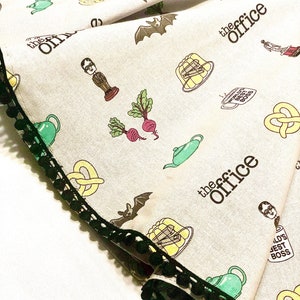 May include: A white tea towel with a black and green trim. The tea towel features a repeating pattern of images and text related to the TV show "The Office". The images include a bat, a teapot, a pretzel, a beet, a character from the show, and a bowl of jelly. The text includes the words "The Office" and "World's Best Boss".