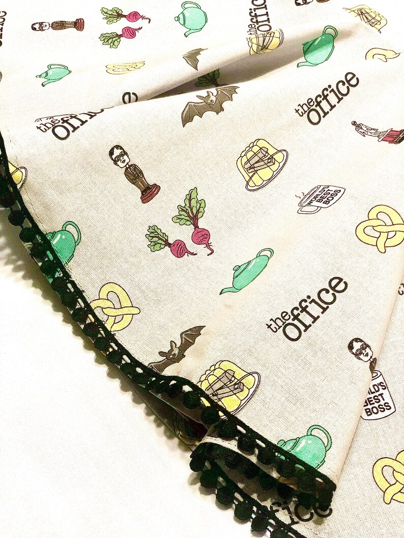 May include: A white tea towel with a black and green trim. The tea towel features a repeating pattern of images and text related to the TV show "The Office". The images include a bat, a teapot, a pretzel, a beet, a character from the show, and a bowl of jelly. The text includes the words "The Office" and "World's Best Boss".
