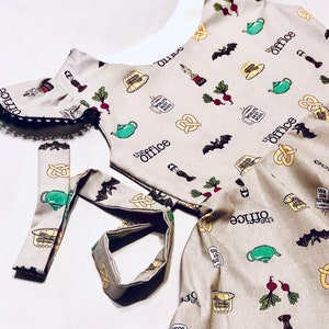 May include: A beige dress with a white collar and short sleeves. The dress is covered in a pattern of The Office characters and objects, including a teapot, a pretzel, a beet, and a bat. The dress has a black lace trim around the neckline and sleeves. The dress also has a matching beige sash.