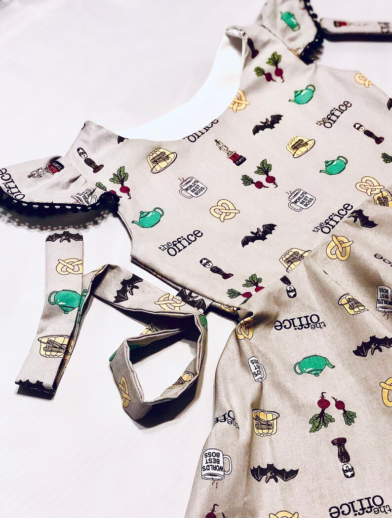 May include: A beige dress with a white collar and short sleeves. The dress is covered in a pattern of The Office characters and objects, including a teapot, a pretzel, a beet, and a bat. The dress has a black lace trim around the neckline and sleeves. The dress also has a matching beige sash.