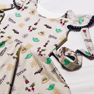 May include: A grey sleeveless dress with a white underlay and black trim. The dress has a print of various images from the TV show "The Office" including a teapot, pretzel, beet, and a mug that says "World's Best Boss".