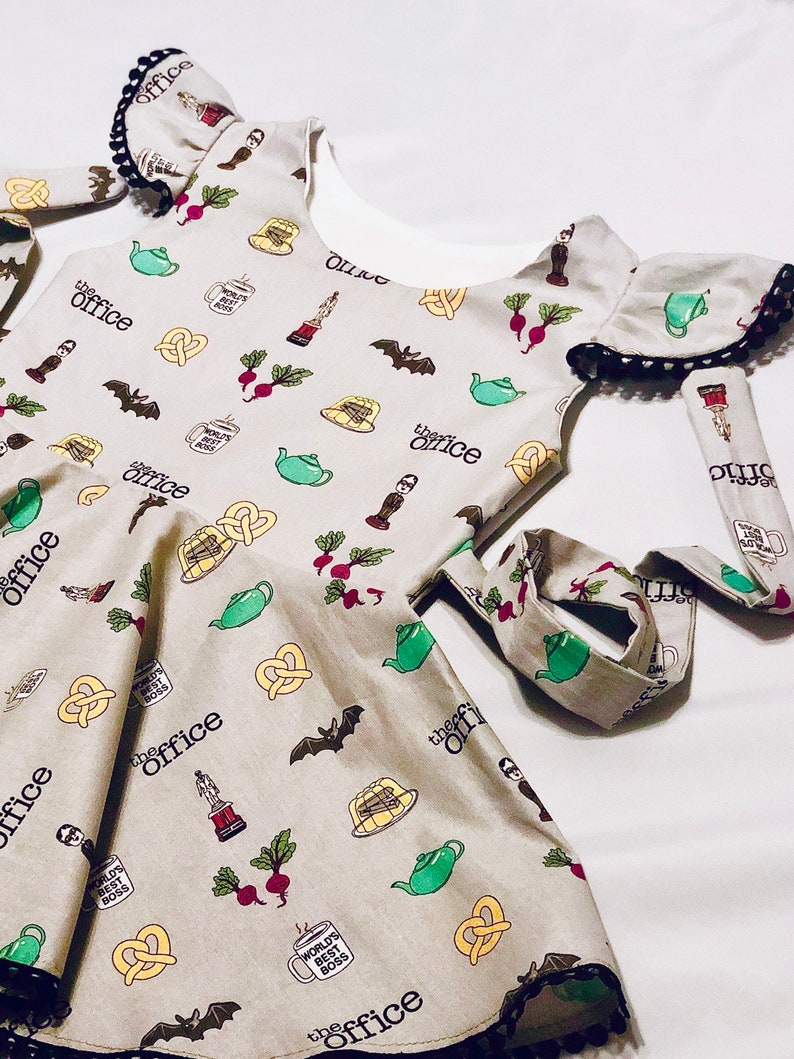 May include: A grey sleeveless dress with a white underlay and black trim. The dress has a print of various images from the TV show "The Office" including a teapot, pretzel, beet, and a mug that says "World's Best Boss".