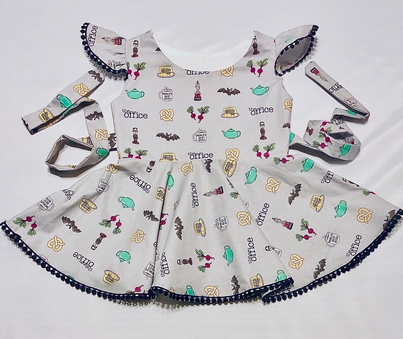 May include: A grey sleeveless dress with a black trim and a white background. The dress is covered in a pattern of The Office characters and objects, including the words "The Office" and "Office".