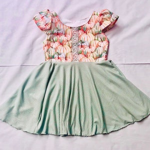 May include: A light green corduroy dress with a pumpkin print bodice. The bodice features a white lace trim and flutter sleeves. The skirt is a solid light green color.