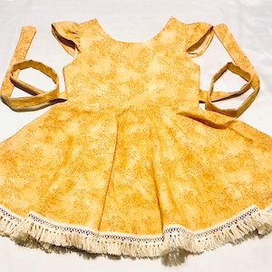 May include: A yellow dress with a floral pattern and white fringe trim. The dress has a scoop neck and short sleeves. It has a tie at the back.