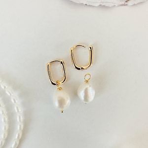 May include: A pair of gold-colored, rectangular hoop earrings with a dangling, white, teardrop-shaped pearl. The earrings are displayed on a white surface, with a pearl necklace and a seashell in the background.