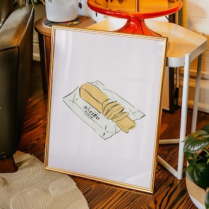 May include: A framed art print with a butter illustration. The artwork shows a stick of butter, partly sliced, on a white wrapper. The frame is gold, and the print is displayed in a room with plants and furniture.