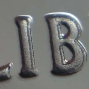 May include: Close-up of a silver coin with the word "LIBE" embossed on it.
