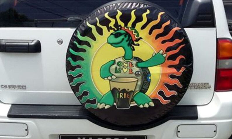 Puede incluir: Un coche blanco con una cubierta de rueda de repuesto que presenta una tortuga verde tocando un tambor. La tortuga lleva gafas de sol y un sombrero. La cubierta de la rueda tiene un dise&ntilde;o de sol amarillo, verde y naranja con las palabras "One Love" e "Irie" en ella.