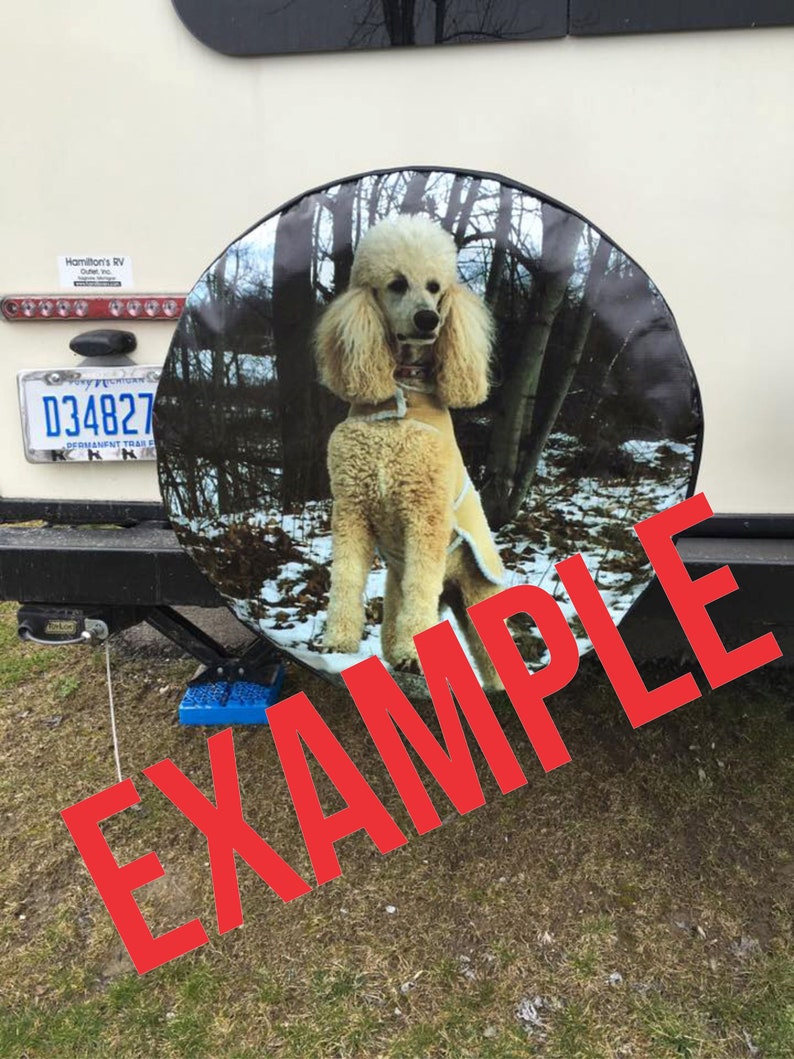 May include: A spare tire cover with a white standard poodle in a snowy forest setting. The cover is black with a white border. The text "EXAMPLE" is superimposed over the image in red.