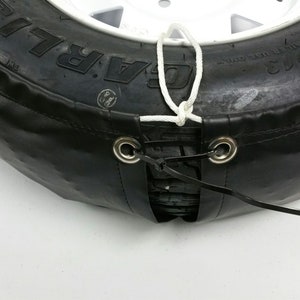 May include: A black tire cover with a white rope tied around it. The cover has two metal grommets and a black strap attached to the grommets.