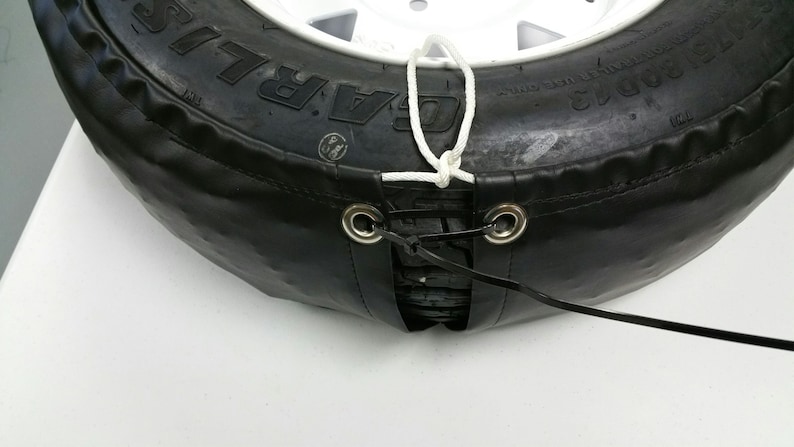 May include: A black tire cover with a white rope tied around it. The cover has two metal grommets and a black strap attached to the grommets.