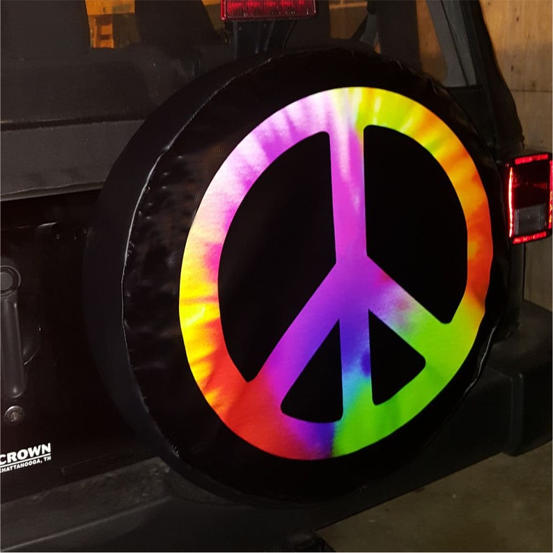 May include: Black spare tire cover featuring a large, colorful peace sign in a tie-dye pattern. The peace symbol is rendered in vibrant hues of yellow, orange, red, purple, and green. The cover is mounted on the back of a black vehicle.