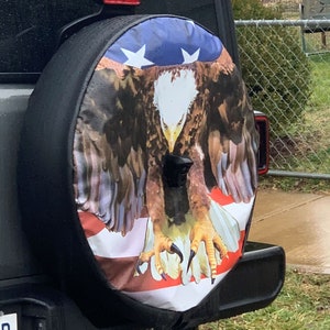 May include: A black spare tire cover with a bald eagle and American flag design. The eagle is perched on the flag with its wings spread out. The flag is red, white, and blue.