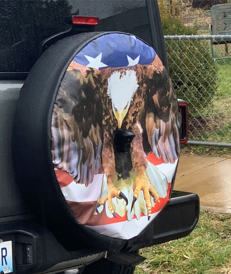May include: A black spare tire cover with a bald eagle and American flag design. The eagle is perched on the flag with its wings spread out. The flag is red, white, and blue.